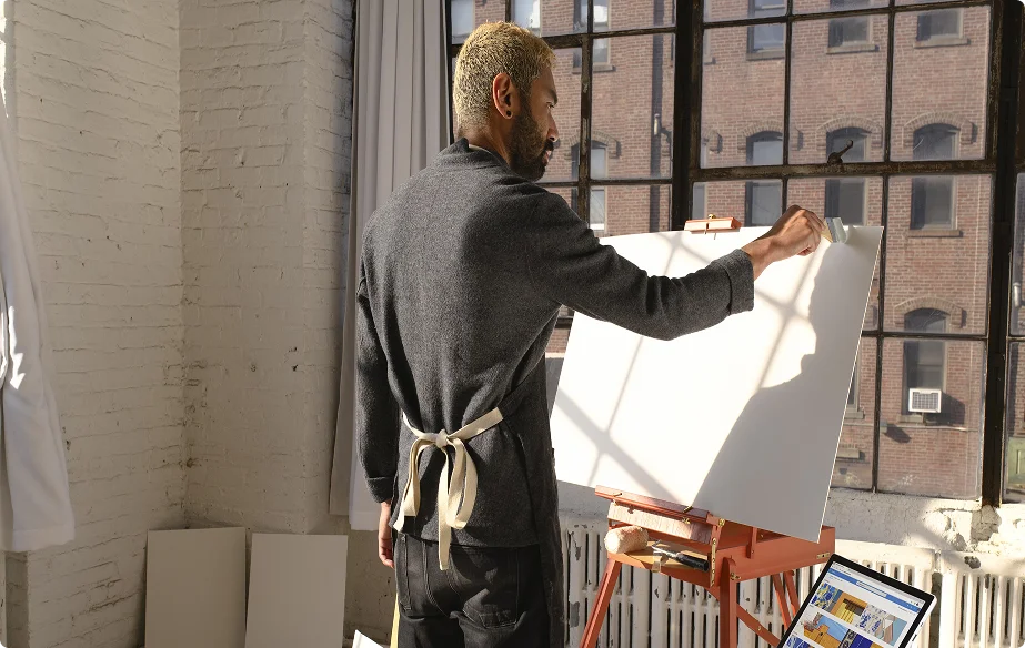 An artist works at an easel beside a sunlit studio window.