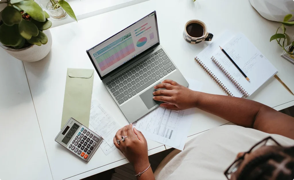 A business owner reviews charts on a laptop beside a notebook, calculator, and coffee.